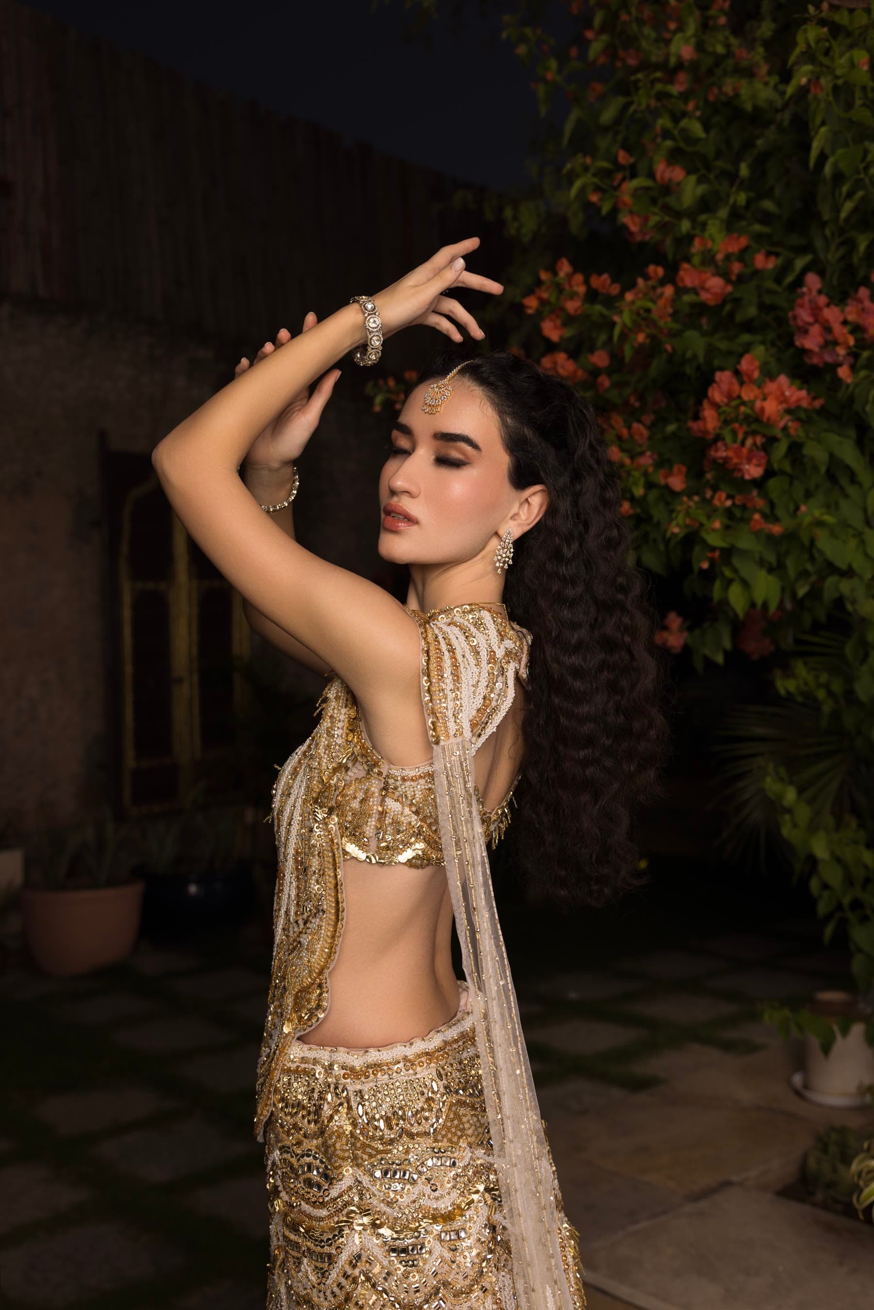 Both arms raised overhead, gold sari against the dark